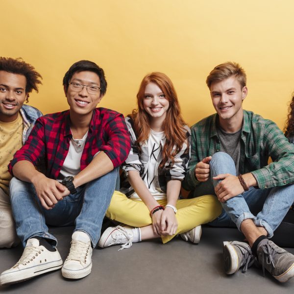 Multiethnic group of smiling young friends sitting on the floor together over yellow background